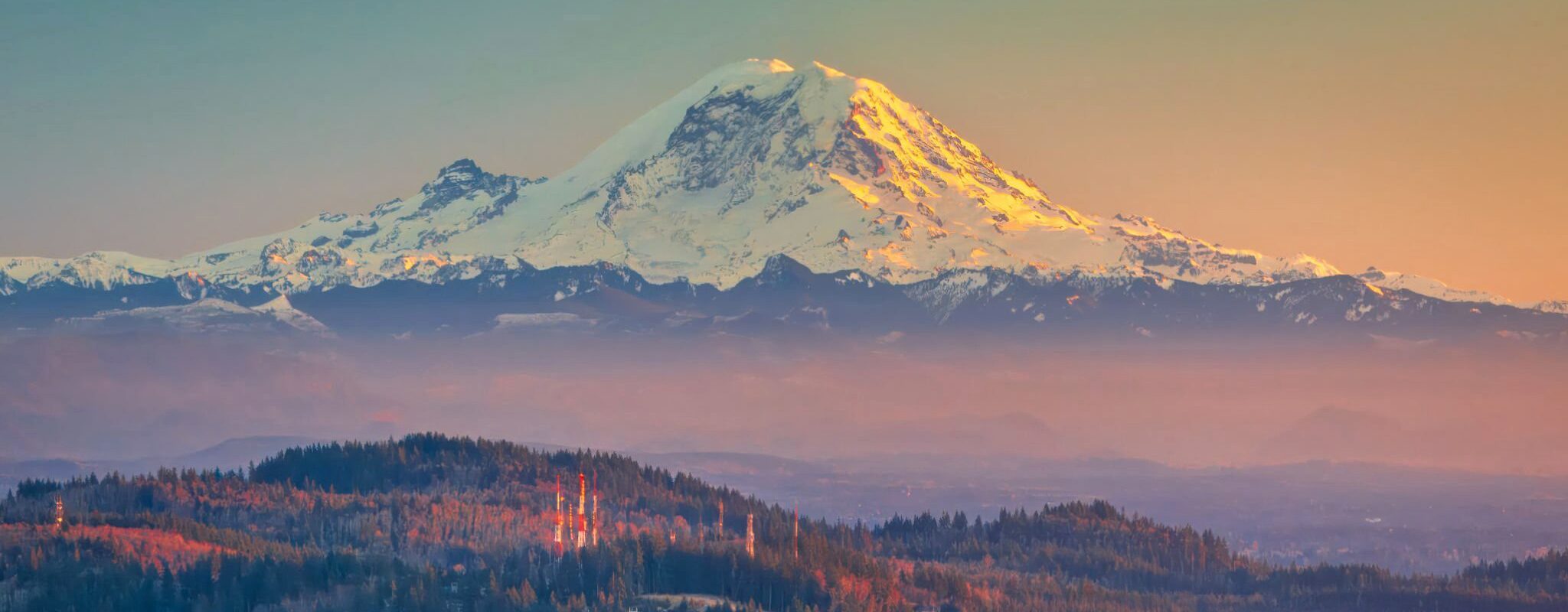 Sunset light illuminating the snow-capped peak of Mount Rainier above rolling forested hills.