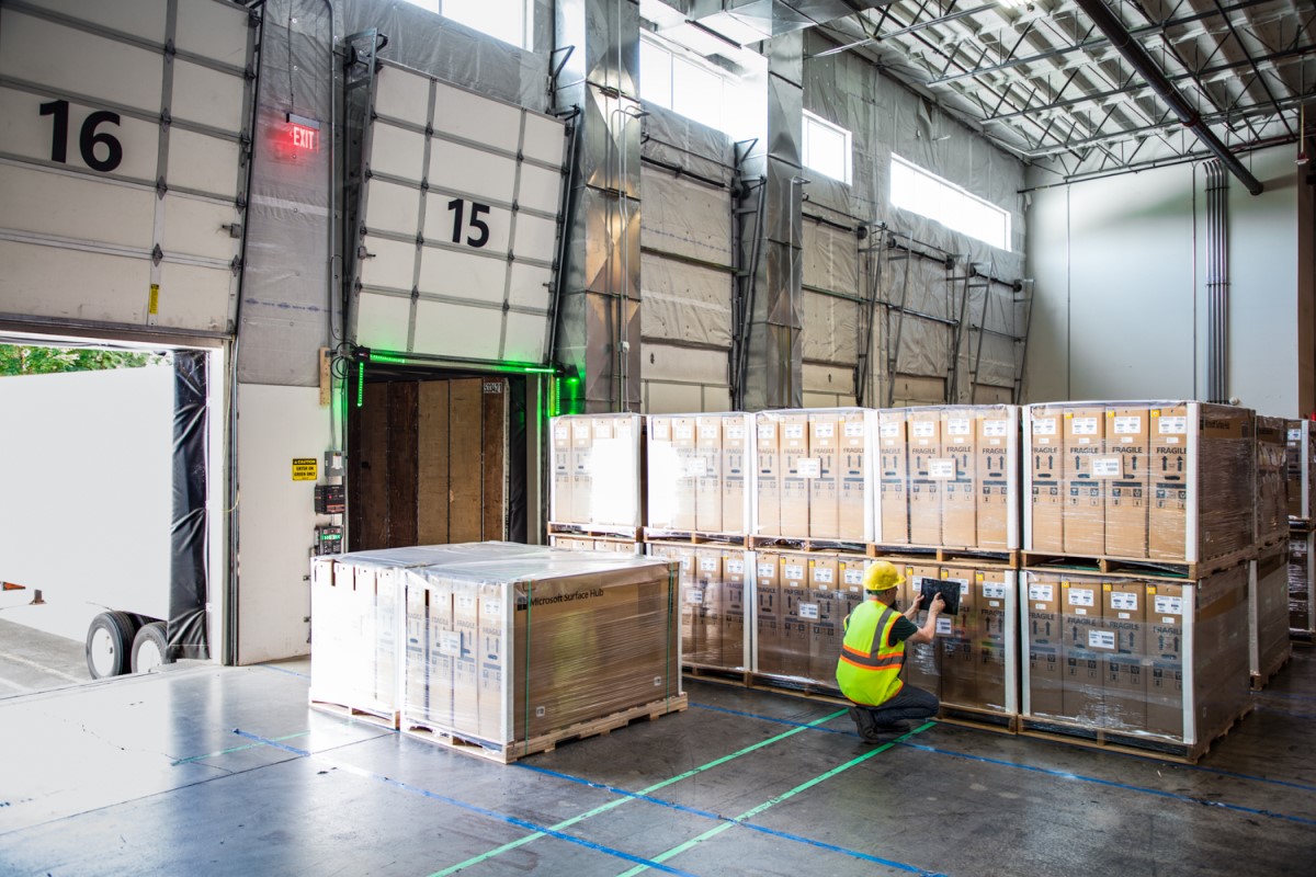 Male worker in hardhat and reflective vest using tablet in warehouse loading dock. Tablet screen not visible