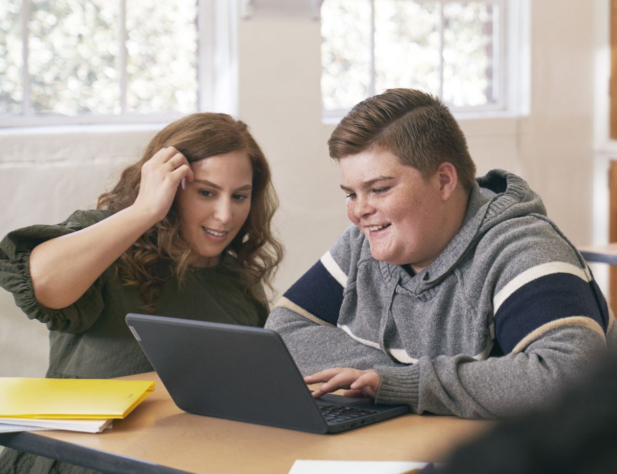 An educator kneeling at a student’s desk and helping him with schoolwork on his laptop