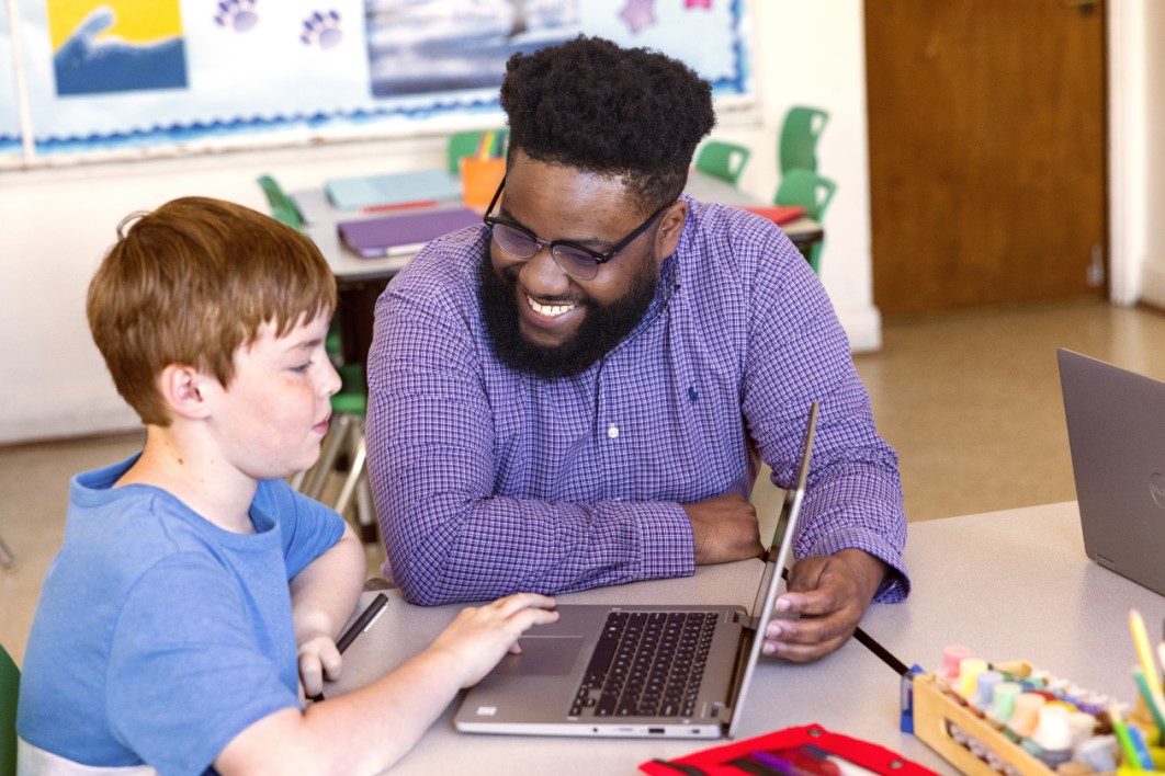 A teacher kneeling at a table engaging with a student who&rsquo;s working on a laptop
