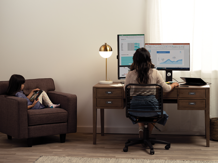 The future of work includes flexibility: A person sitting in a living room, at a desk working. A child is sitting on the sofa next to it.