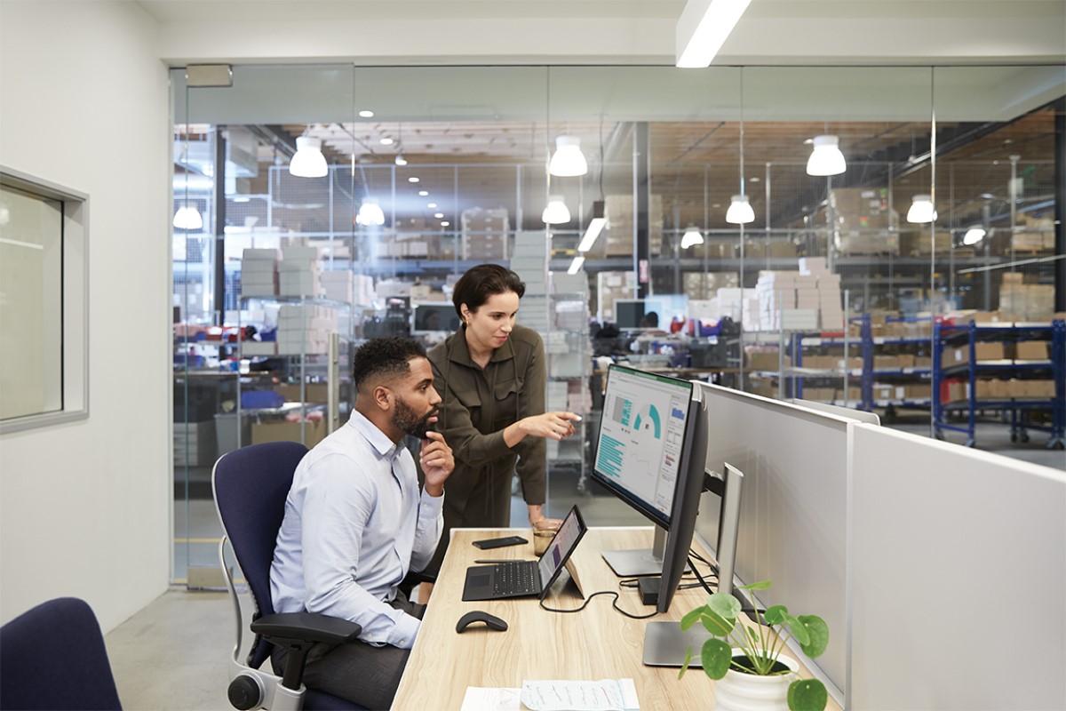 Two adults in an office adjacent to a warehouse setting collaborating while overlooking dual monitors