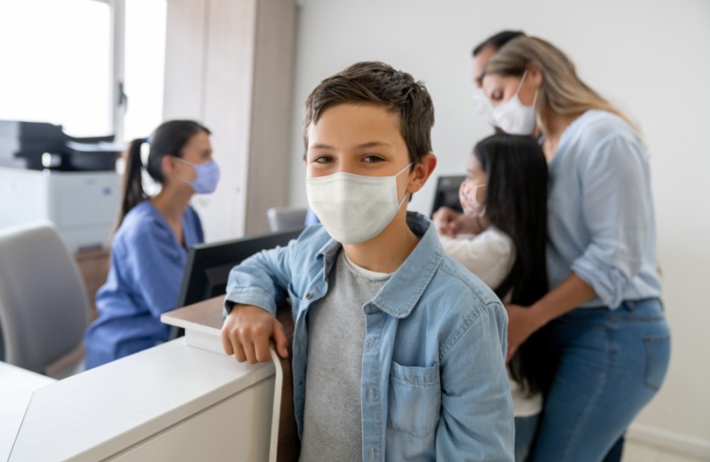 Photo of a young patient with a mask on at looking happy in the foreground. The rest of the family in the background check-in with the office manager