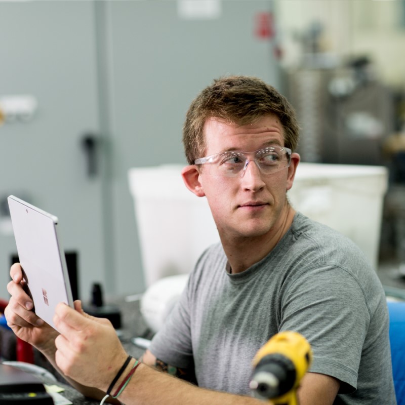 Male factory worker sitting at desk in manufacturing plant. He is wearing safety glasses and looking over his left shoulder while holding a Surface Pro (screen not shown)
