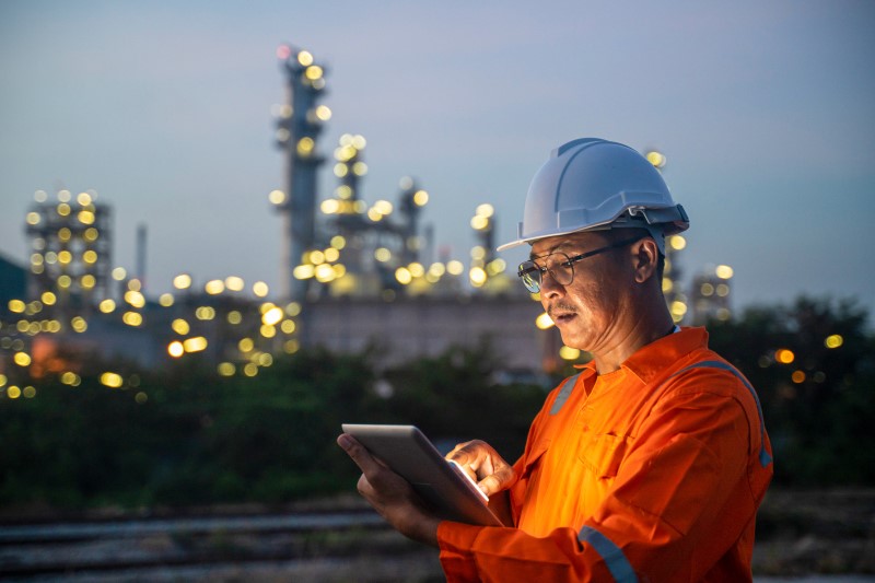 An engineer wearing a hardhat uses a digital tablet while working the night shift at petroleum oil refinery