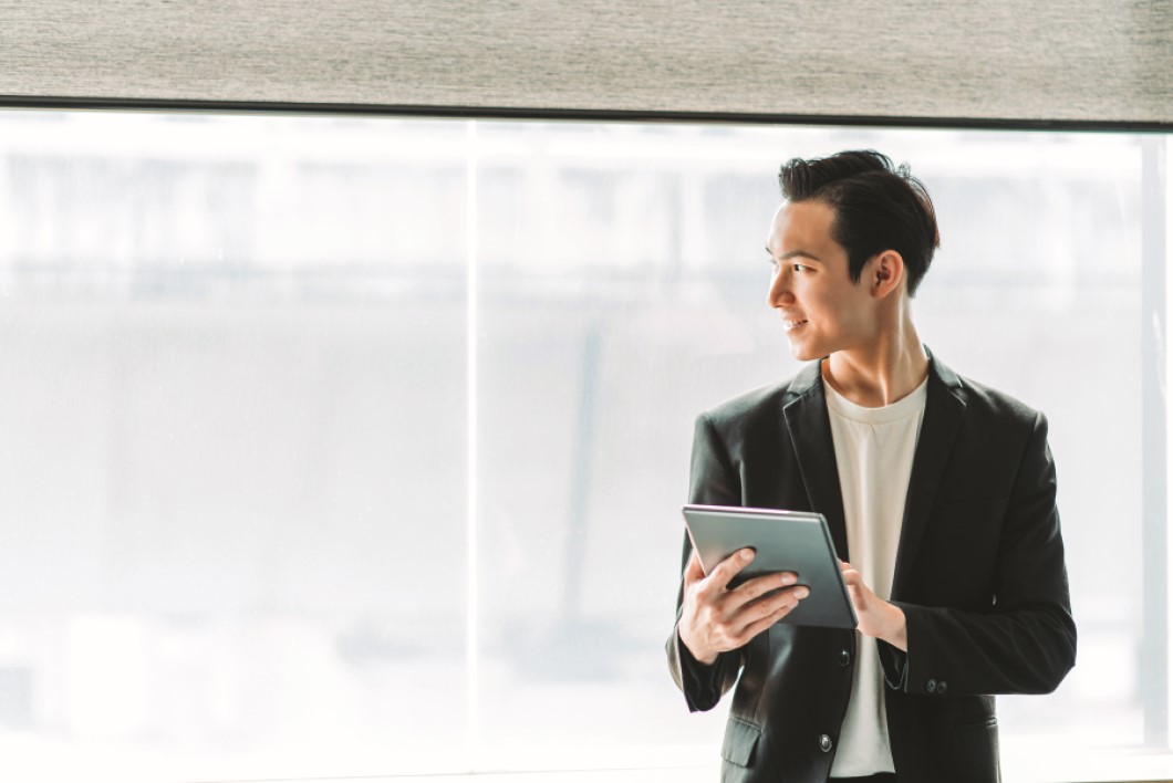An young businessman holding a digital tablet stands in front of a large window in his modern office