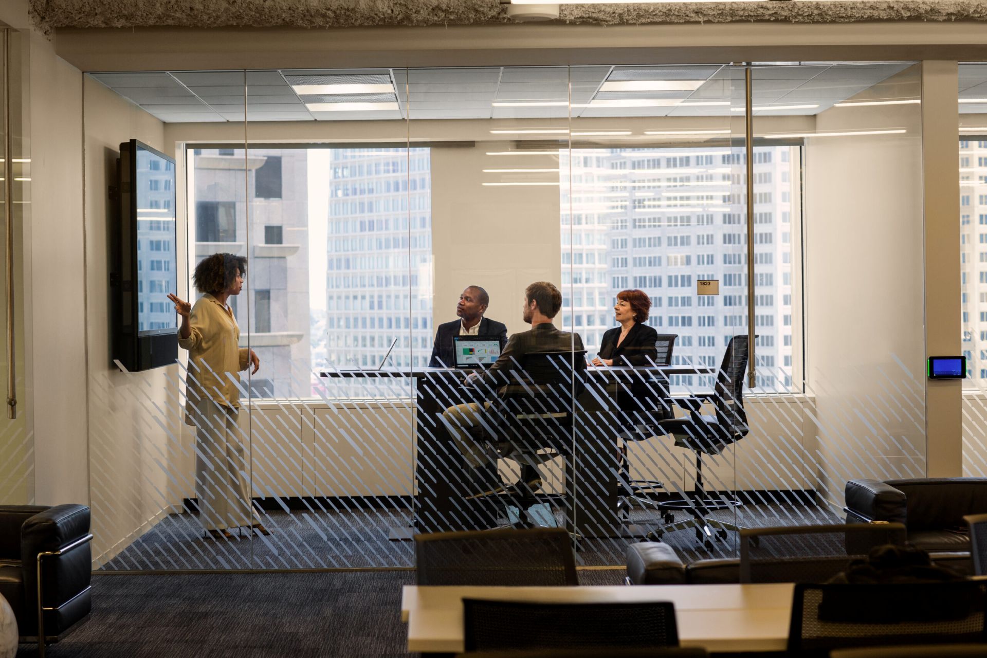 People sitting in a meeting room with one person standing and presenting next to a large screen on the wall