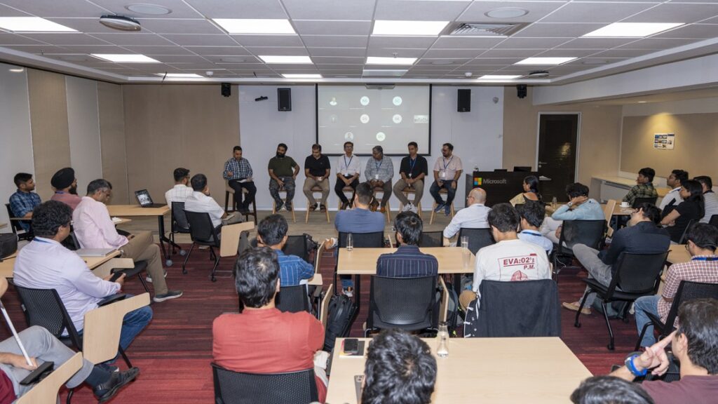 Photograph of a panel discussion in a conference room with six speakers seated on high chairs at the front and an audience of around 25 people seated at tables facing them. The setting includes a projector screen displaying a presentation, a Microsoft podium, and attendees engaged in listening and note-taking.