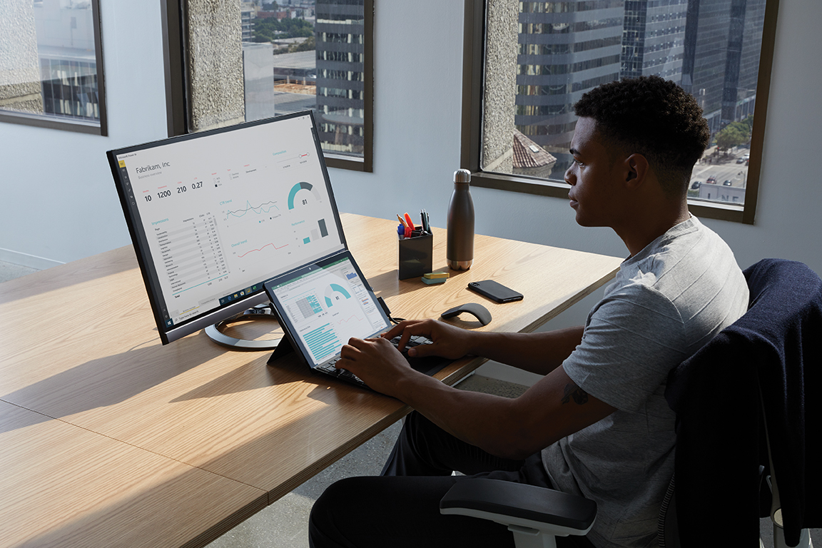 Adult male in an office setting sitting at a desk with his hand poised over the keyboard of a black Microsoft Surface Pro 7 in laptop mode. Microsoft Excel visible on labptop and Microsoft PowerBI screen seen on monitor