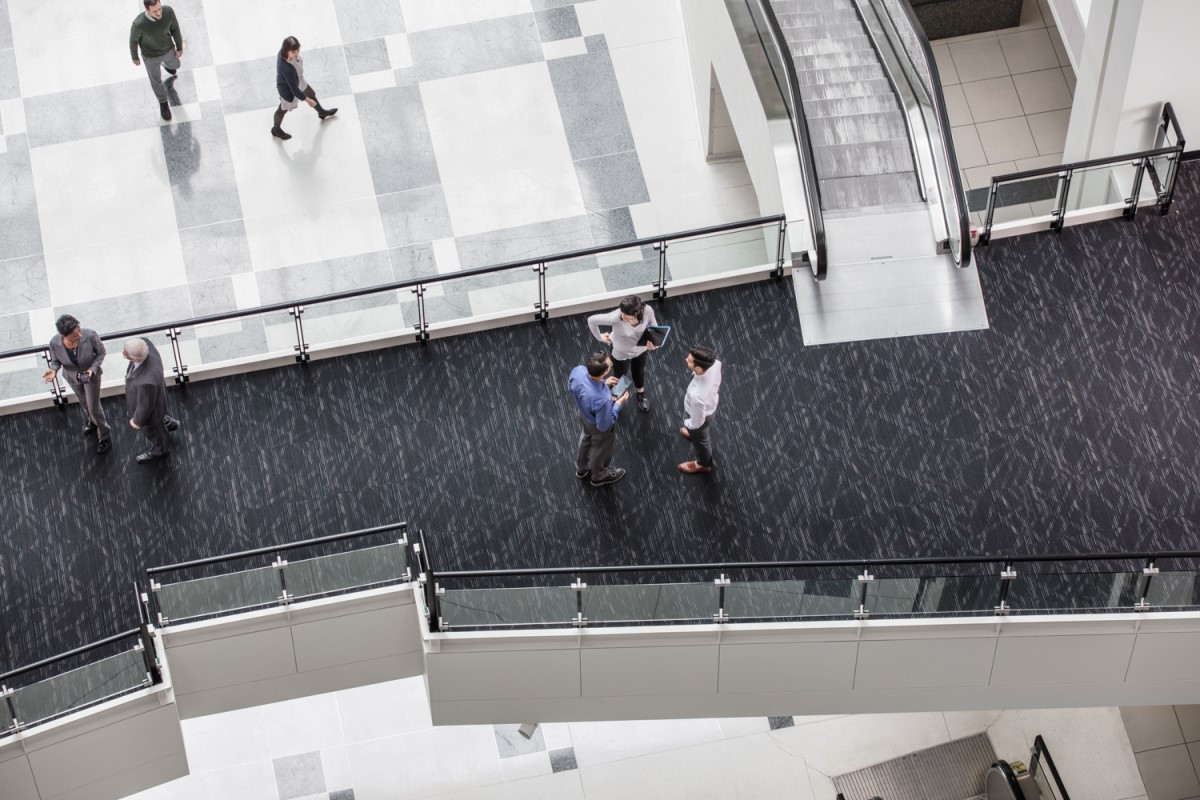 Aerial view of office building&lsquo;s interior entrance hall. Close-up of two separate groups of people standing together on an elevated walkway, chatting. Two others are walking on ground floor below