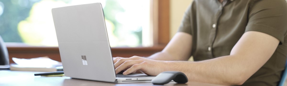 Male teacher sitting at a desk typing on a Surface Laptop Studio in laptop mode. A Microsoft Arc Mouse sits on the desk
