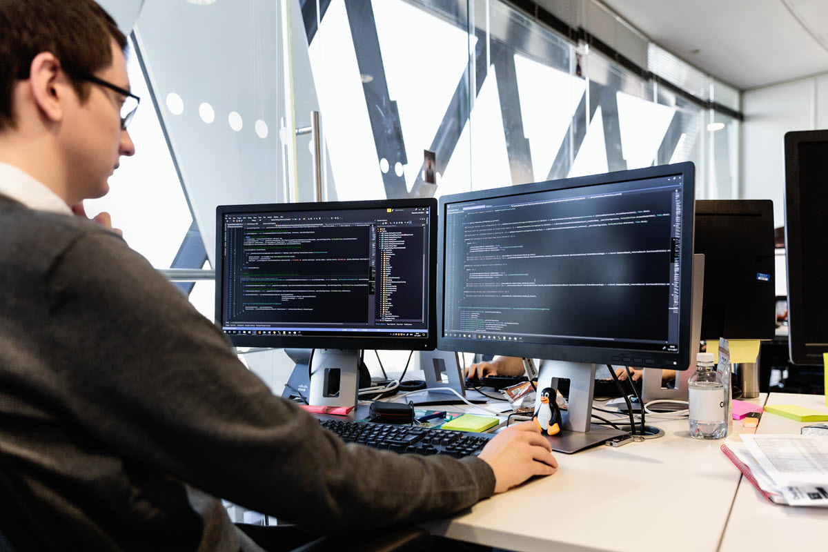 Male developer coding in front of two monitors at desk in office. Programming code shown on both monitors
