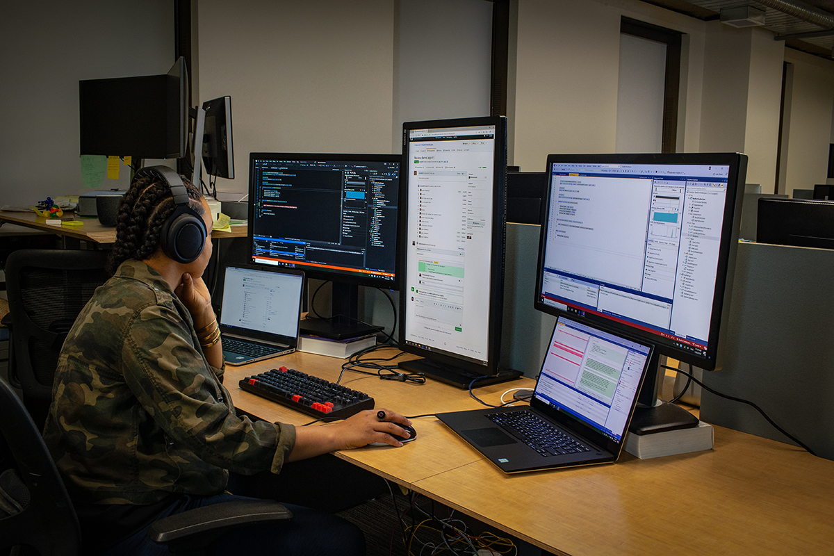 female developer wearing headphones; coding at her PC workspace in an enterprise office, using Visual Studio on a multi-monitor set up