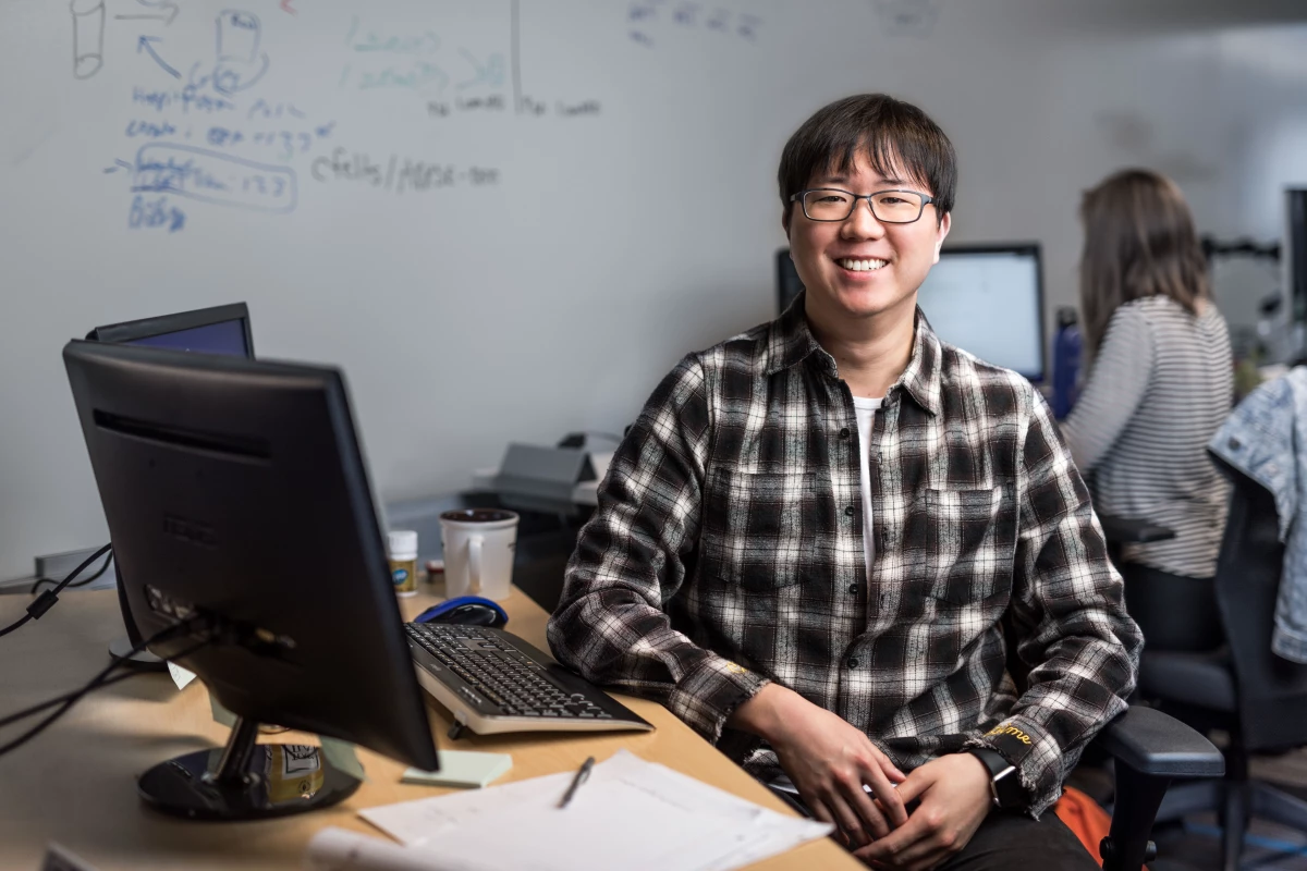 Portrait of male developer in glasses and plaid flannel shirt, smiling and facing camera. He is sitting at a desk in an office setting, with both hands on his lap. Keyboard and desktop computer visible, though screens are not shown. A female worker is shown seated at desk using computer in background (screen blurred). Whiteboard wall also shown in background with handwritten diagrams and formulas