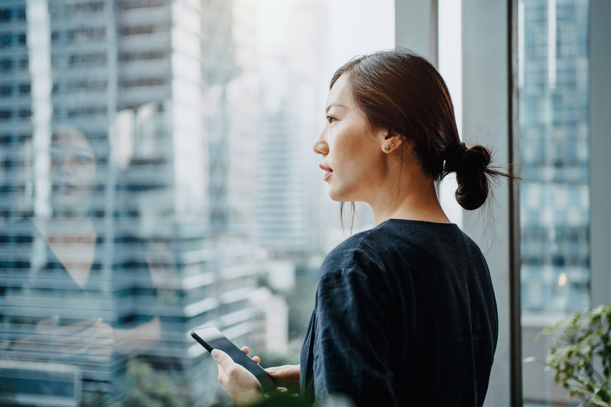 Young businesswoman holding a phone, looking out her office window overlooking the city