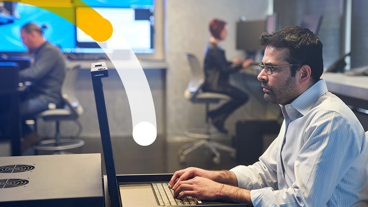 Man in a collared shirt working on a server station inside a secure room. Coworkers and large monitors are in the background