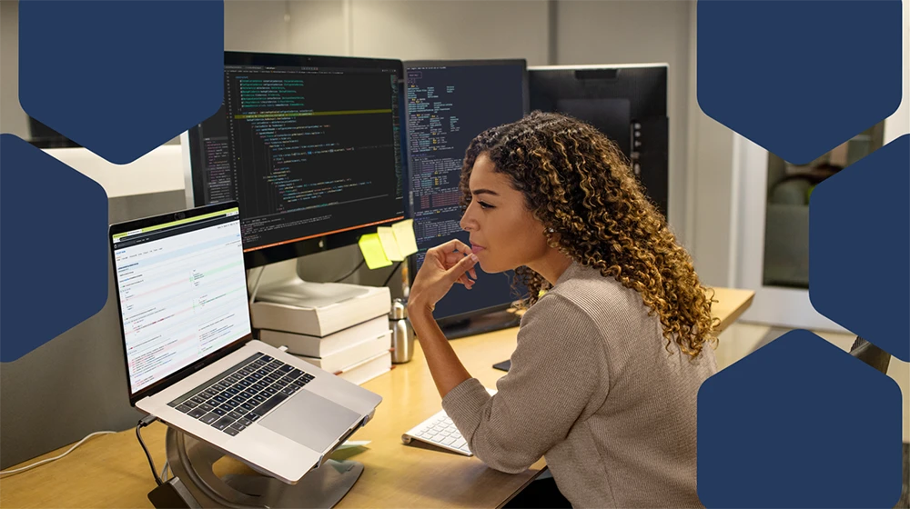 Photo of a female developer working at enterprise office workspace. Focused work. She has customized her workspace with a multi-monitor set up